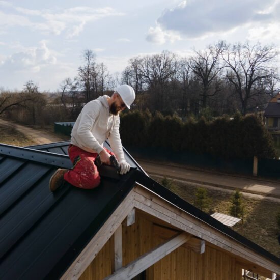 Roofer installing dark metal roof panels on wooden-framed house under construction.