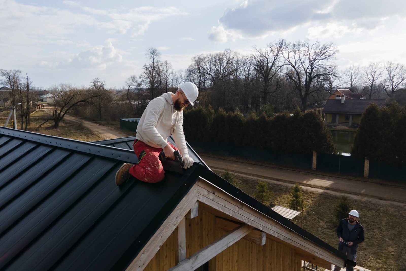 Roofer installing dark metal roof panels on wooden-framed house under construction.