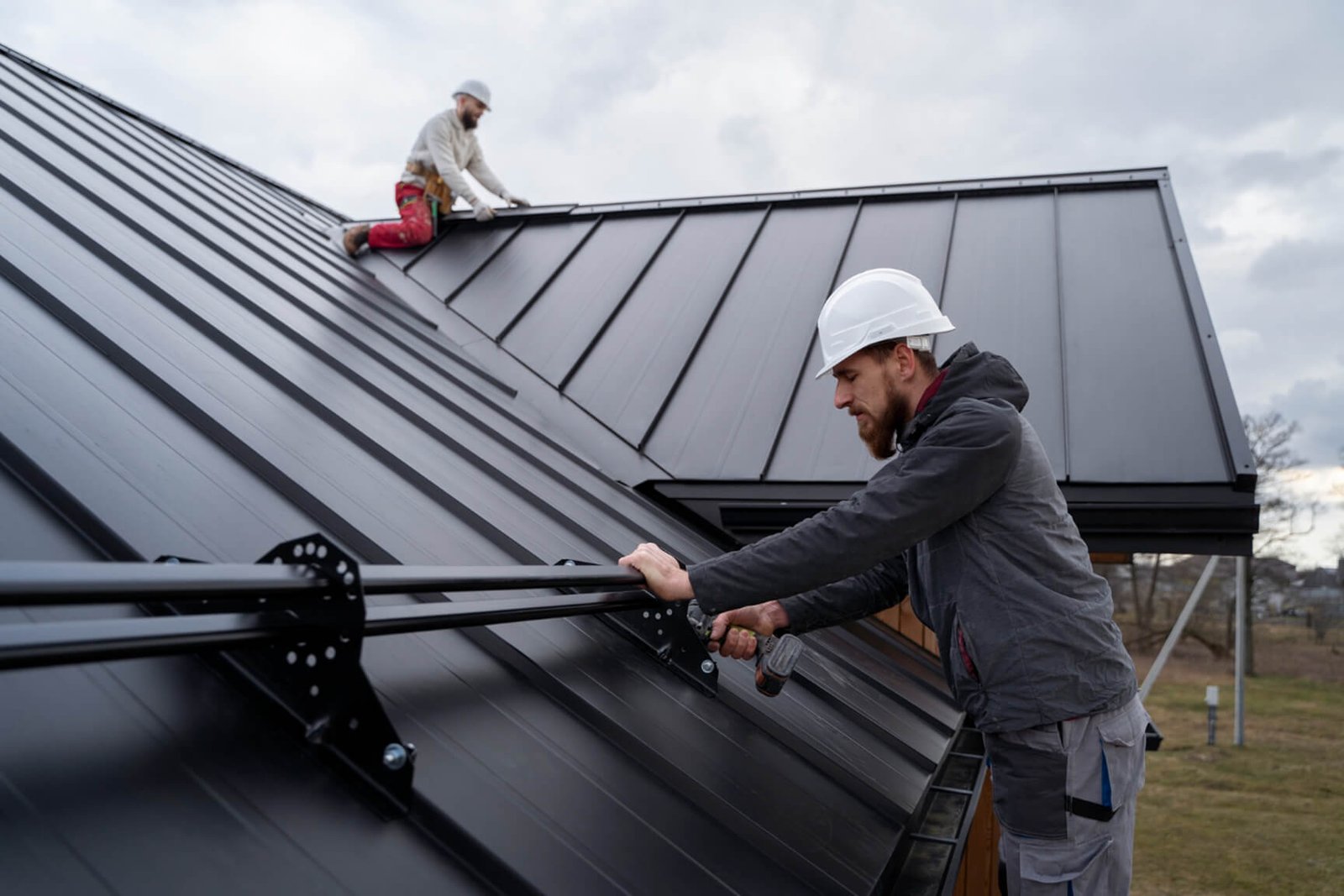 Two roofers installing dark metal roof panels on a residential house.